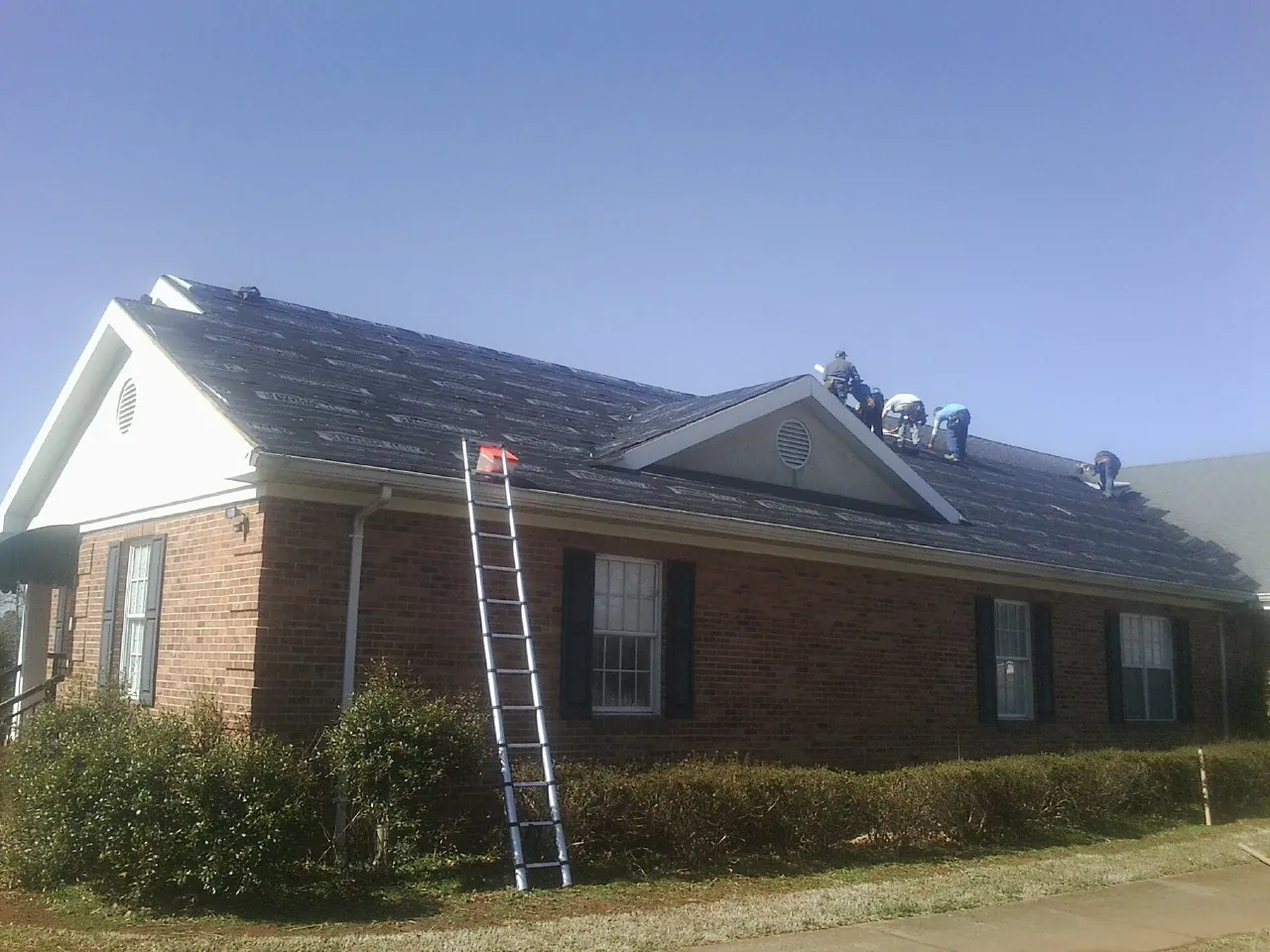 Crew tearing shingles off a brick ranch roof with a ladder set — Emergency Roof Repair work by our crew in Rantoul