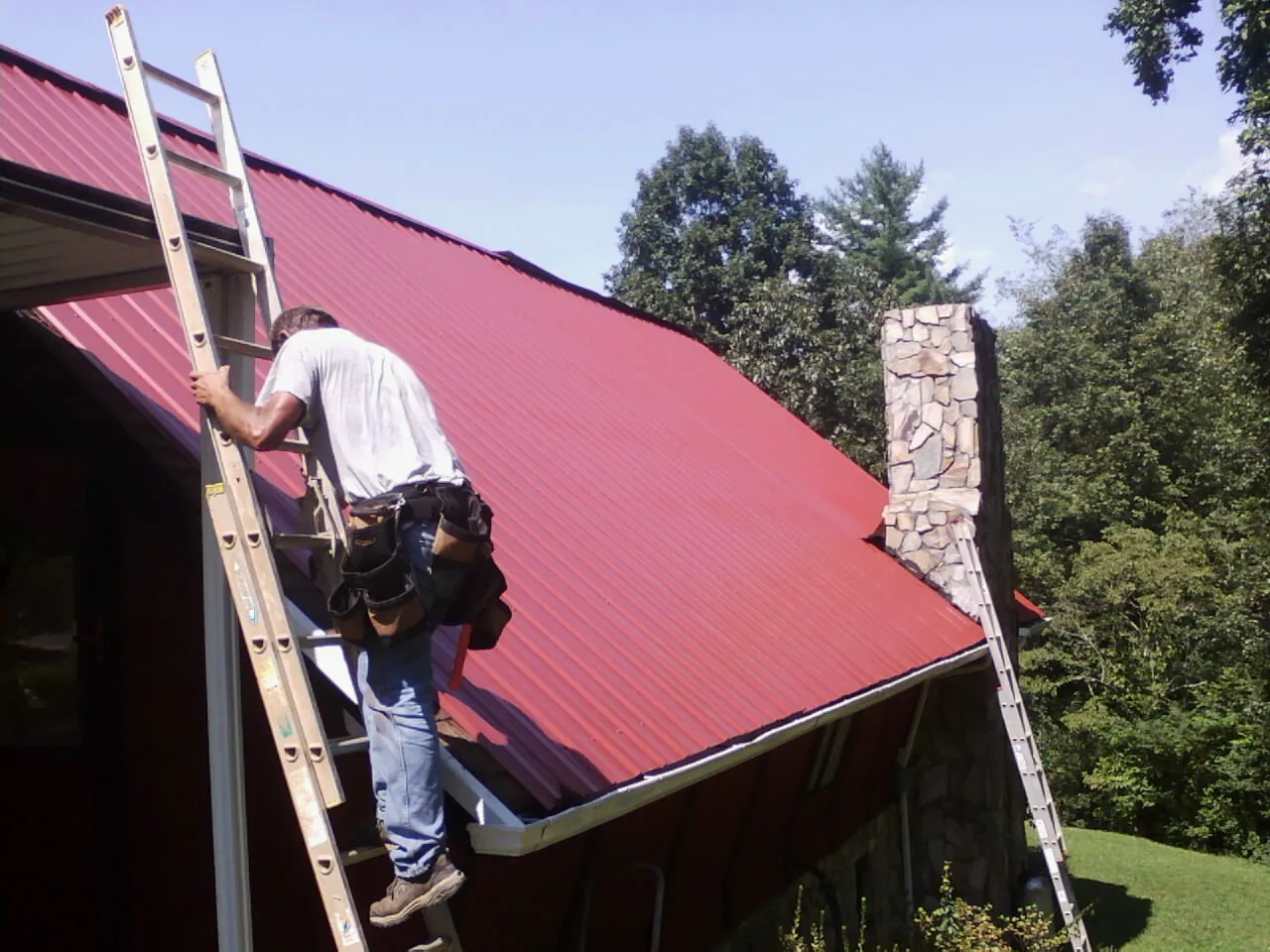 Roofer on a ladder beside a red metal roof and stone chimney — Soffit & Fascia Repair by Roofing Brothers in Rantoul